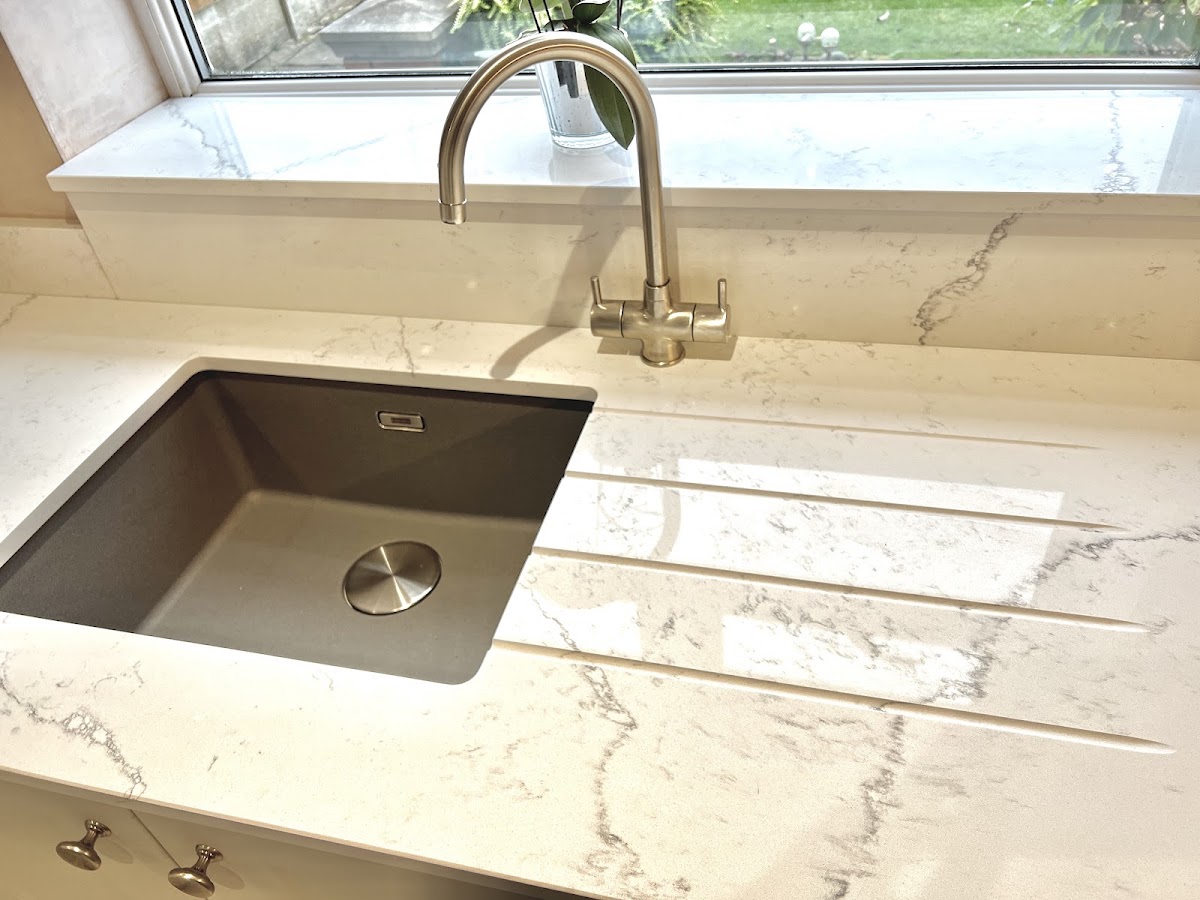 A close-up of a white quartz worktop with a moulded drainer, an undermount composite sink and a brushed-steel mixer tap over the garden window.